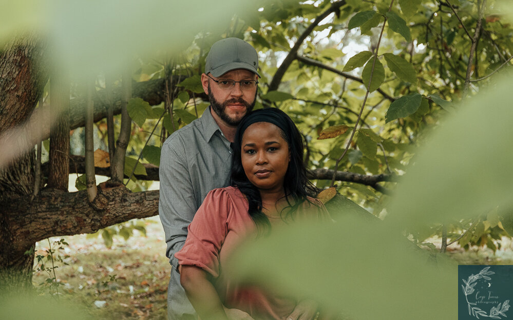 newly engaged couple posing for a photo under a green tree. both looking straight at camera.