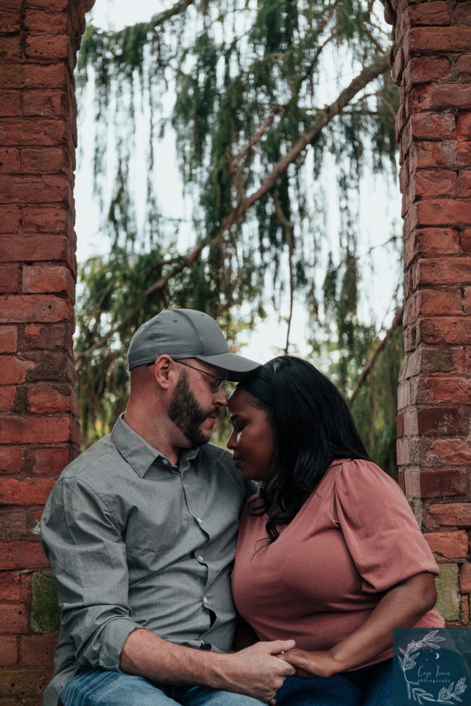 newly engaged couple sitting foreheads touching under brick archway. Engagement Session at Vanderbilt Mansion