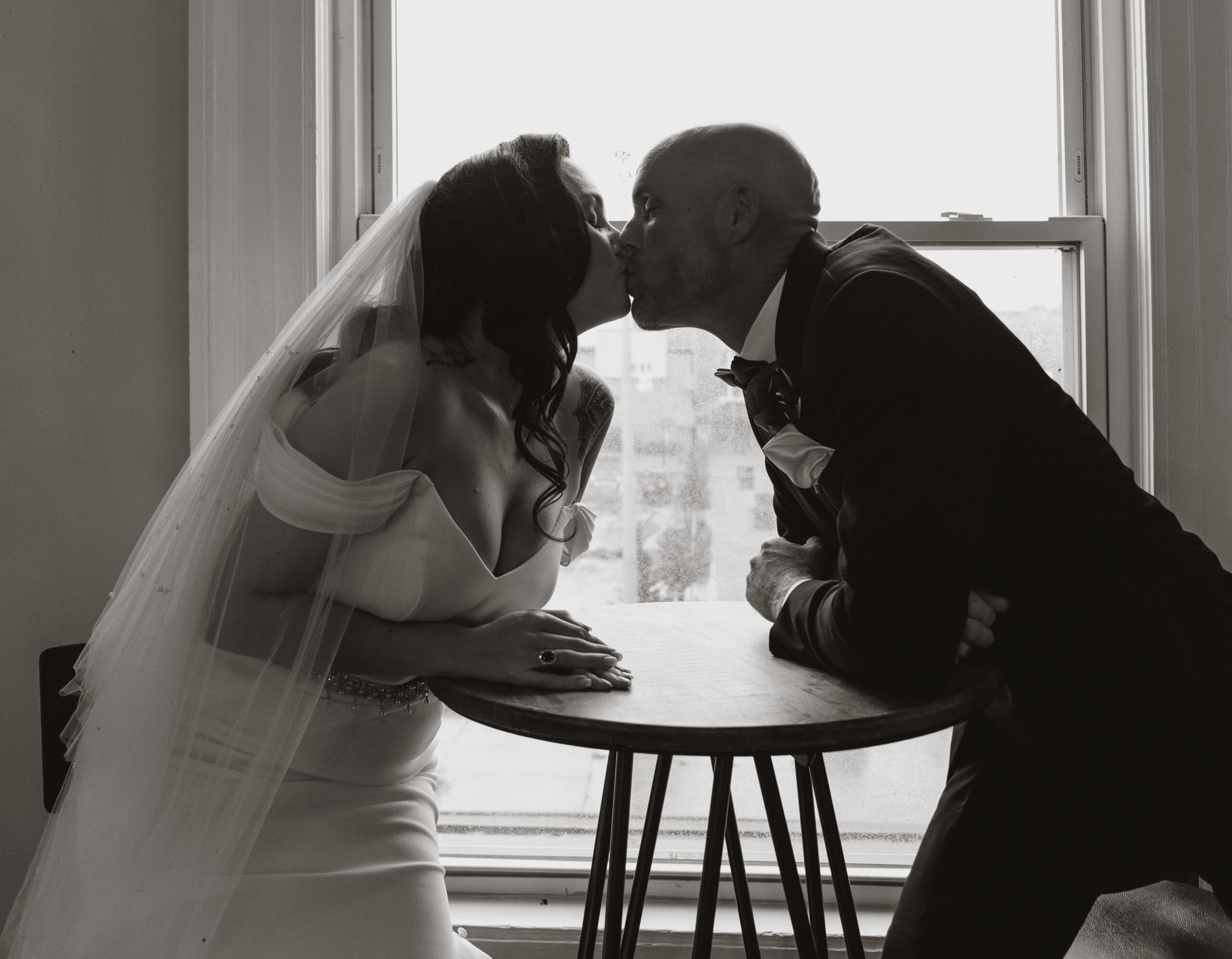 black and white photo of couple kissing in front of window.